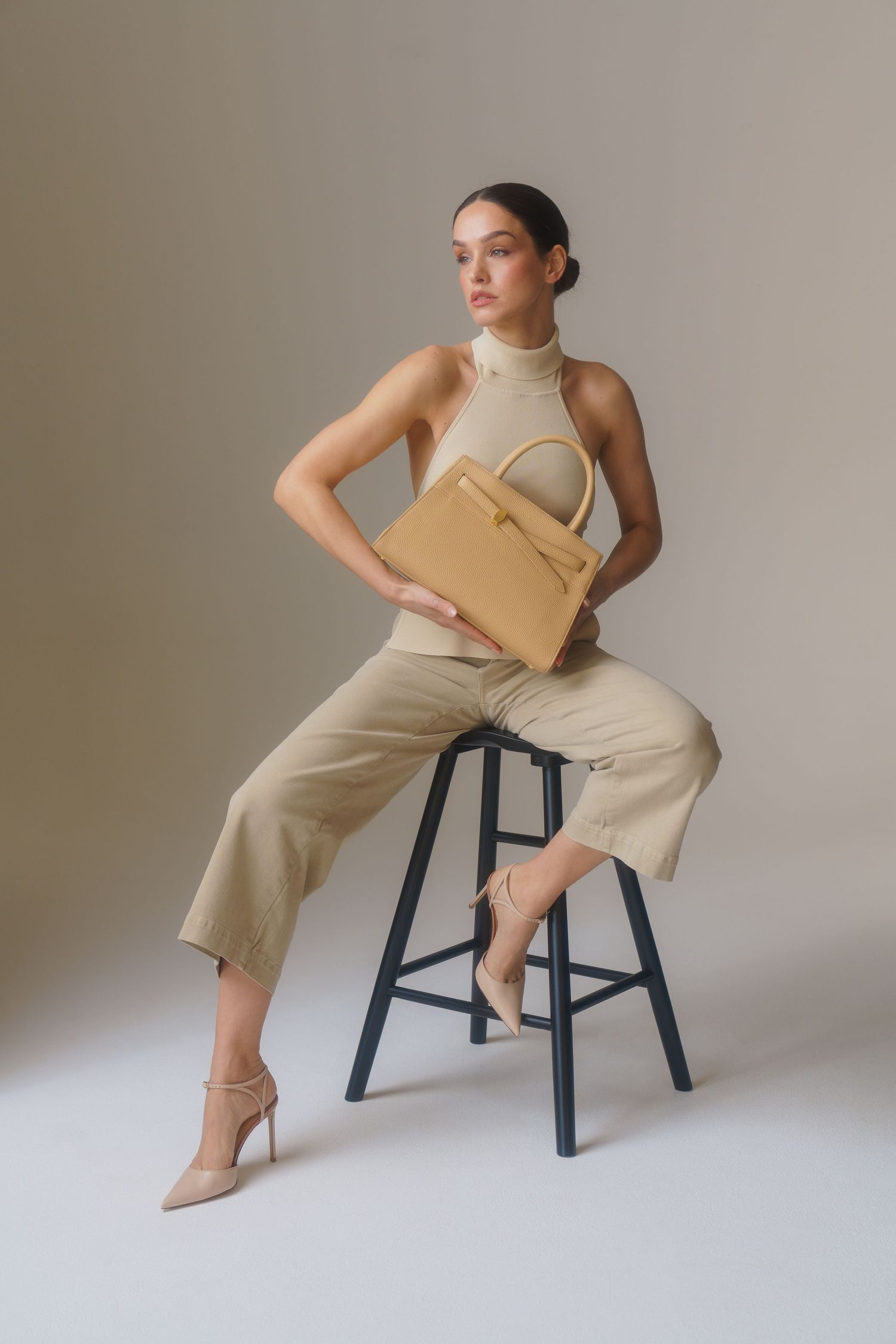 Fashion model in neutral tones poses on stool with tan handbag in minimalist studio setting.