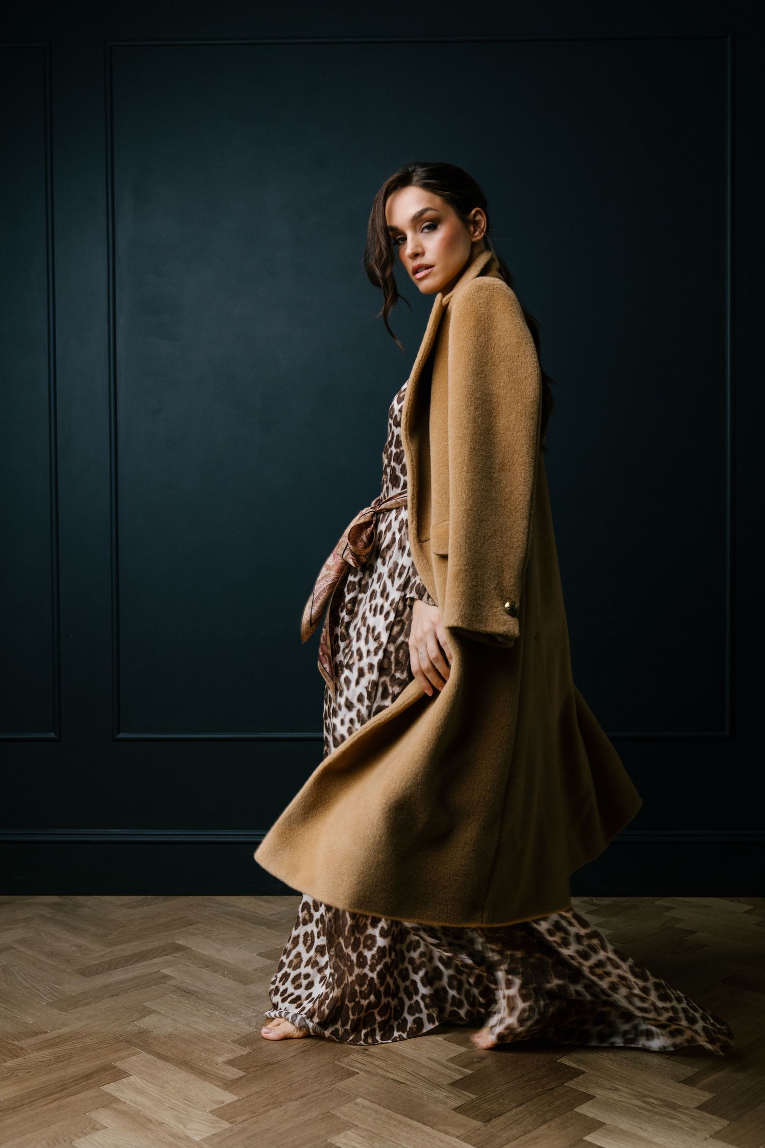 Dramatic fashion portrait featuring camel coat over leopard print maxi dress against dark wall.