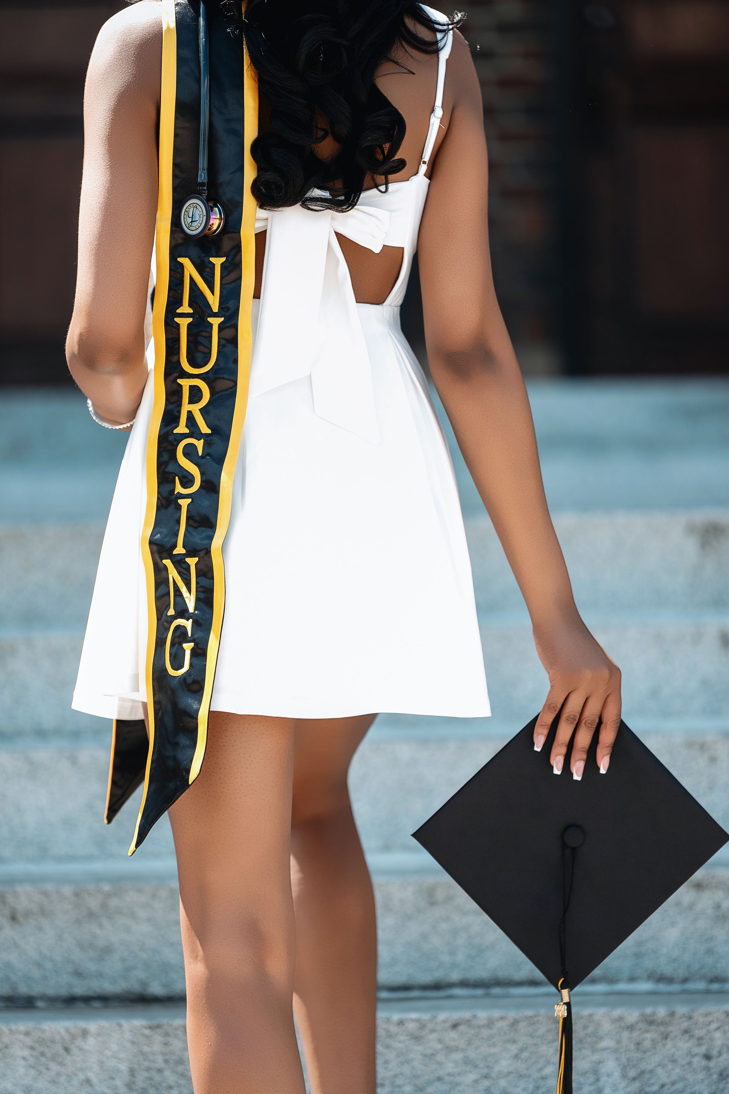 Nursing graduate in white dress holding black mortarboard cap, wearing gold and black nursing graduation sash.