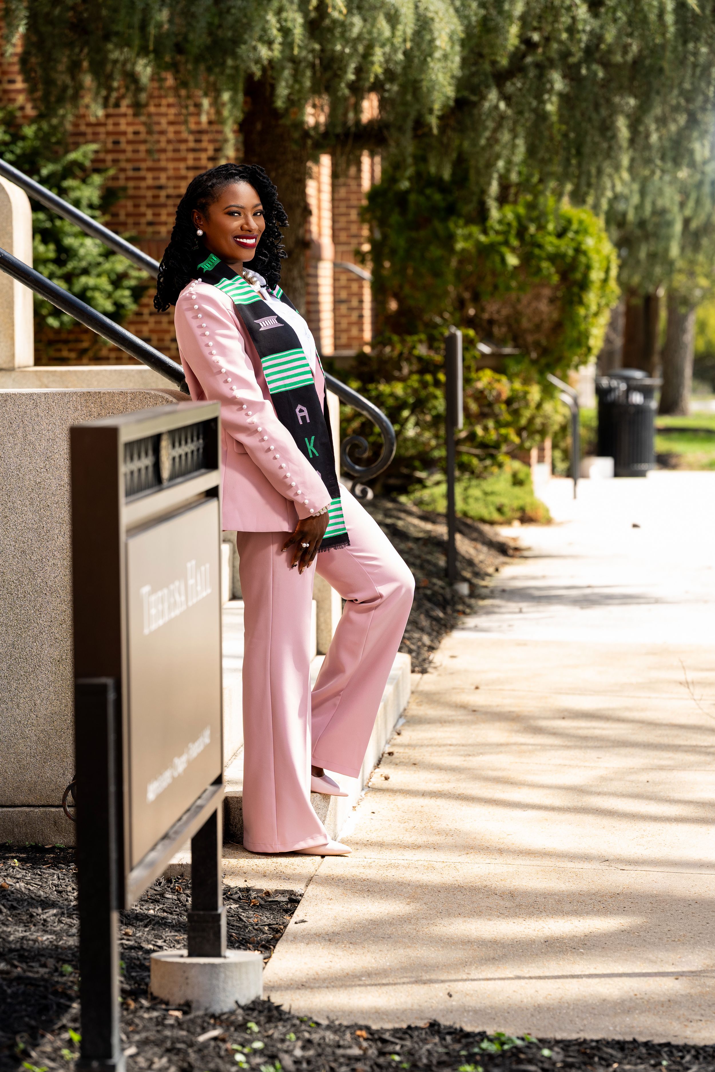 Young Black woman in pink suit with graduation stole smiles on campus steps near Tuskegee Hall sign.