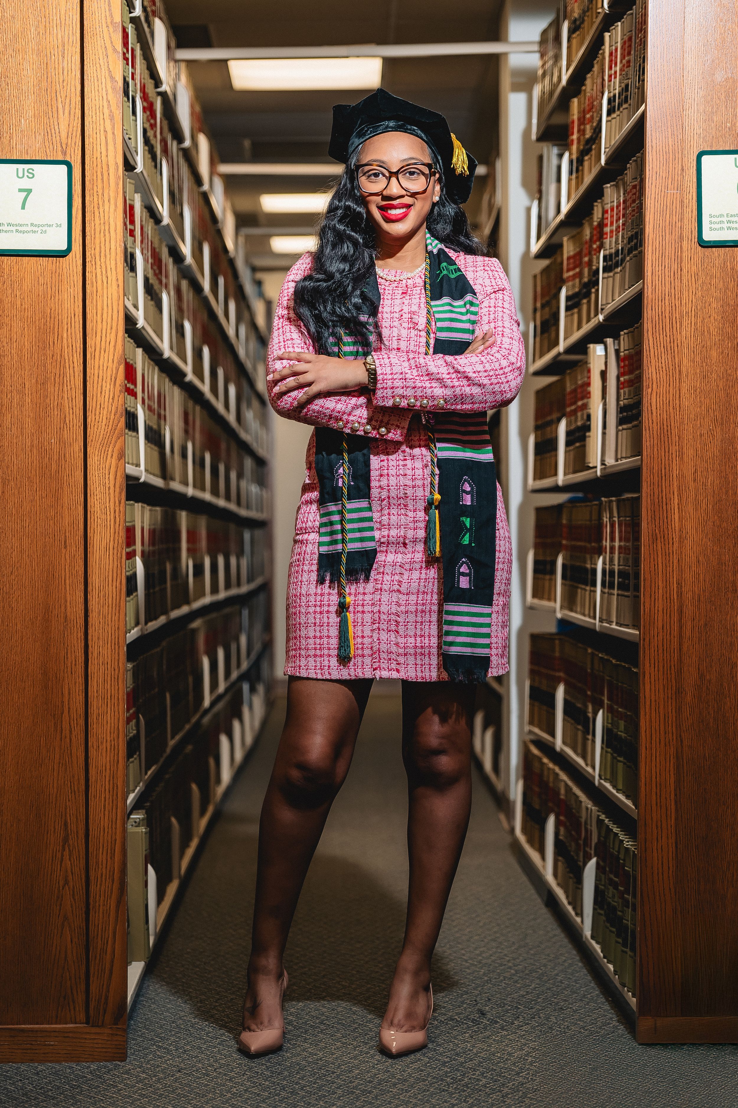 A woman in a pink tweed dress, graduation cap, and kente scarf poses confidently in a library aisle.