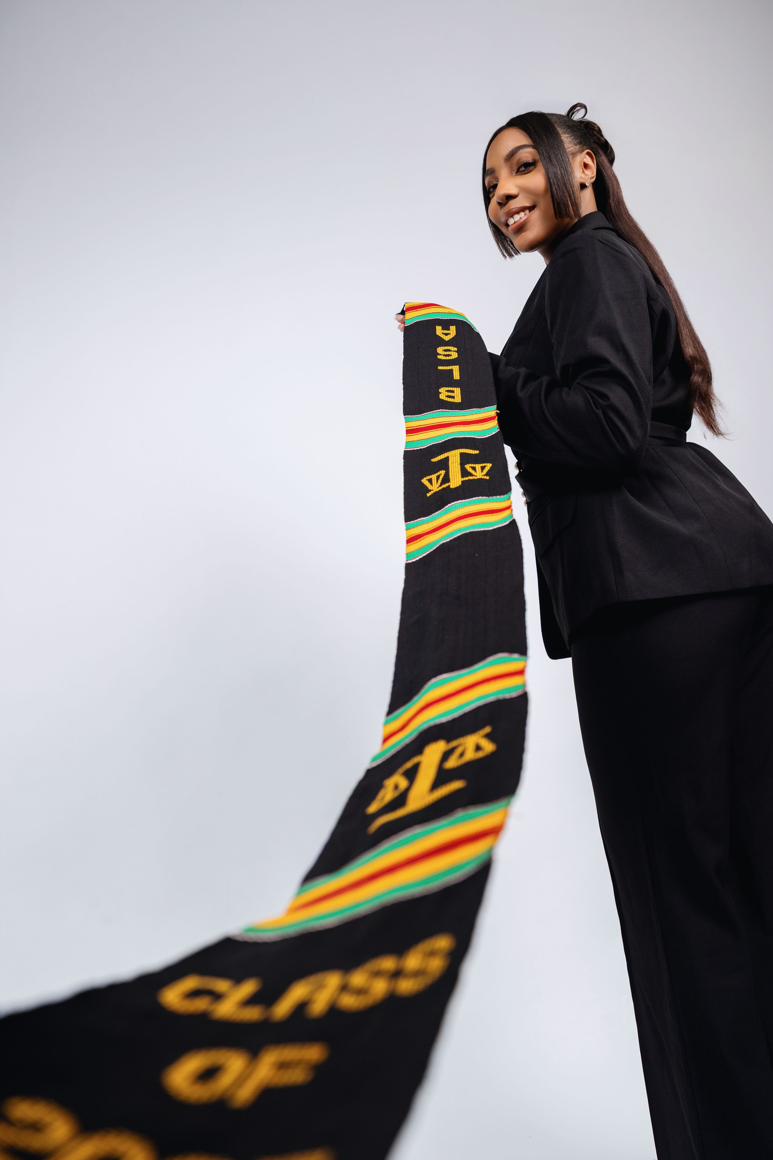 Young woman in black suit holding a graduation kente stole reading Class Of, celebrating academic achievement.