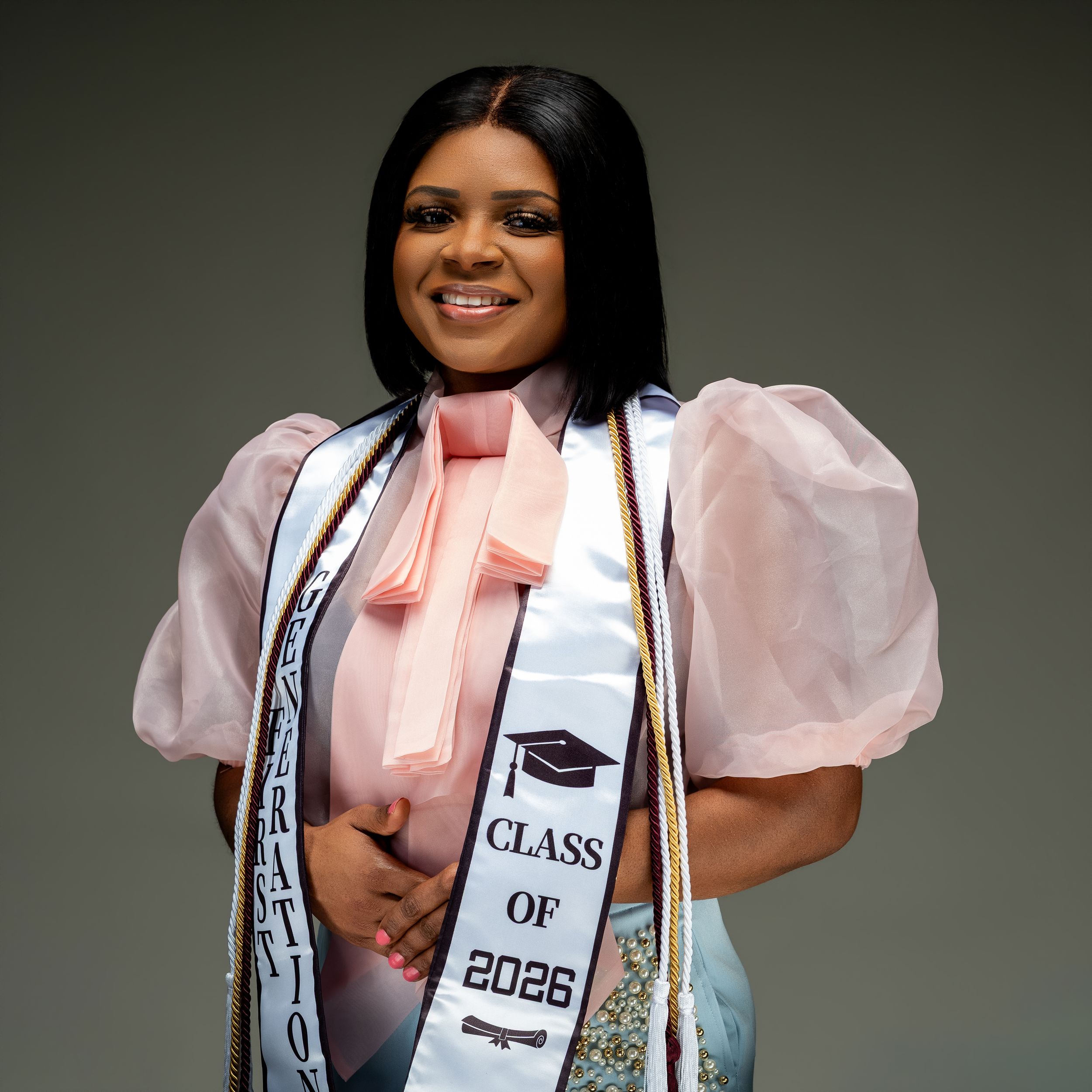 Smiling graduate wearing a Class of 2026 sash and pink organza top poses for a professional graduation photo.