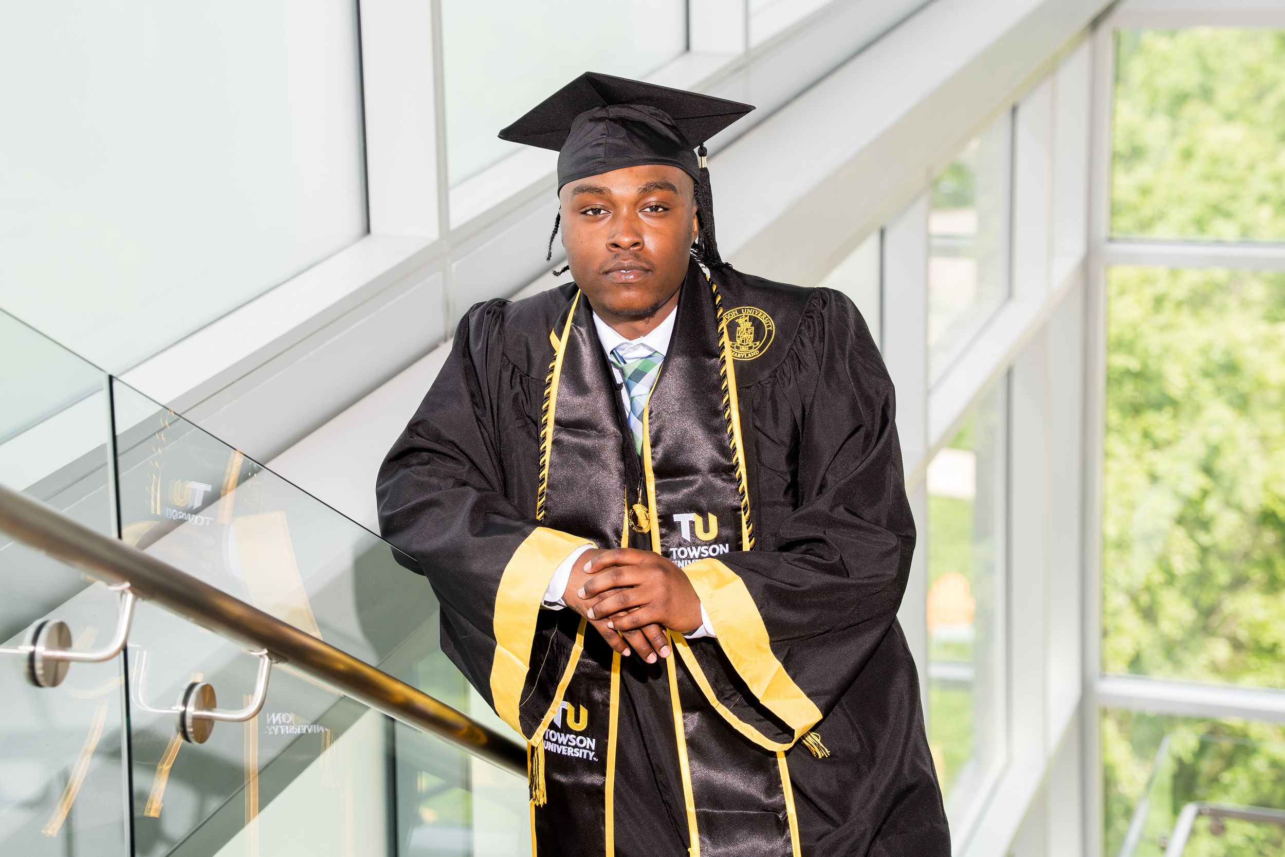 Graduate in black cap and gown with gold stole poses proudly on staircase at Tuskegee University graduation.
