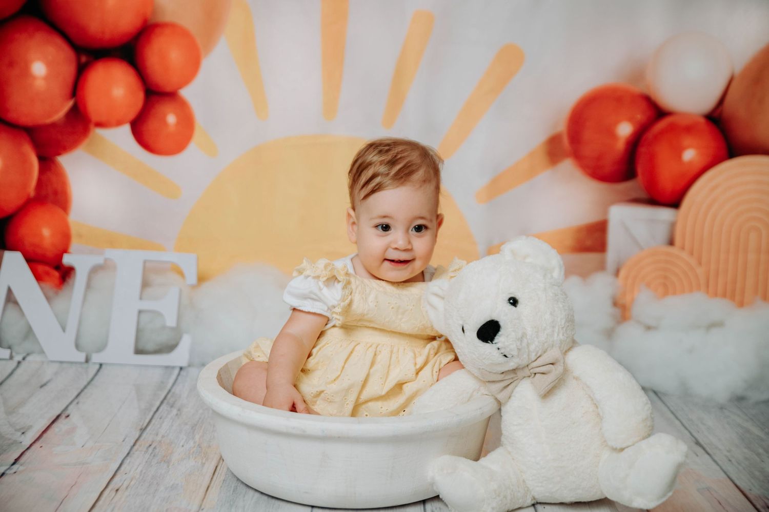 White teddy bear next to basin prop against orange balloon and sunburst backdrop.