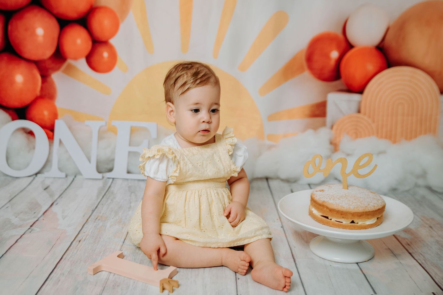 Baby in yellow dress sits on wooden floor with orange balloons and cake for first birthday photoshoot.