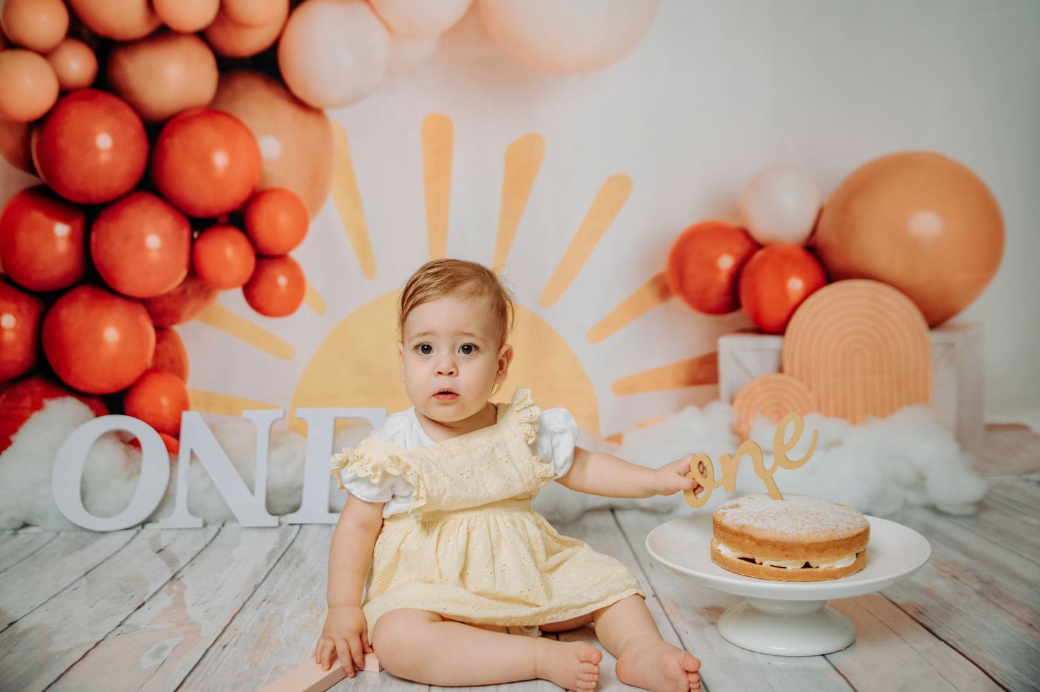 First birthday setup with baby in cream dress reaching toward cake with orange balloon backdrop.