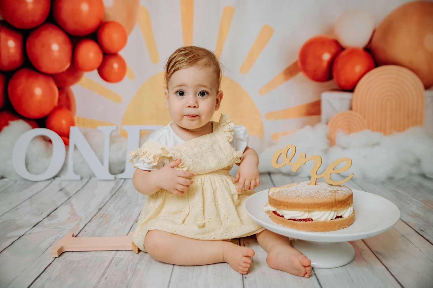 Baby wearing cream dress poses next to birthday cake with orange balloon and sunburst decorations.