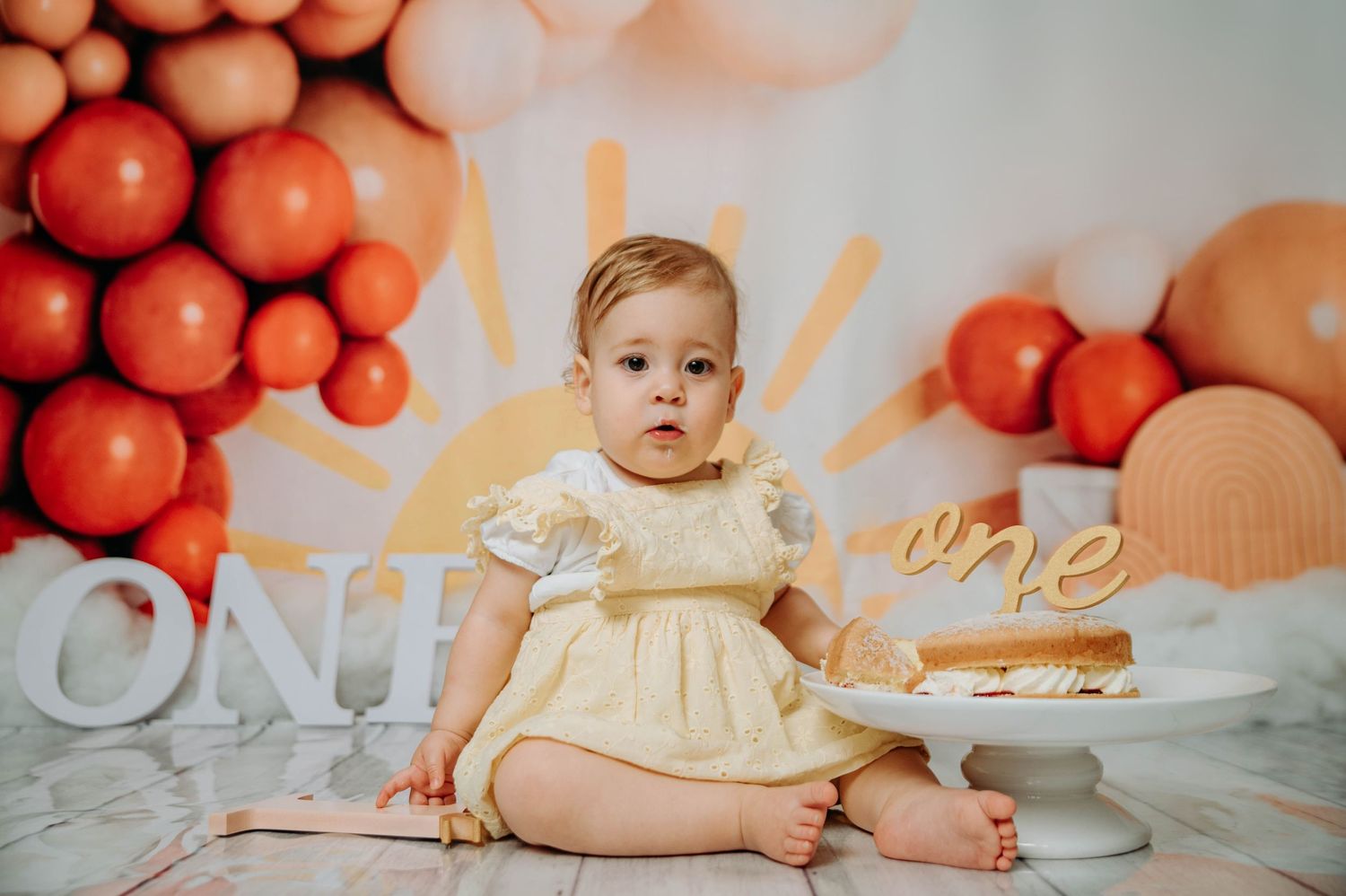 One year old in yellow dress sitting beside cake with orange balloon and sunburst wall decor.