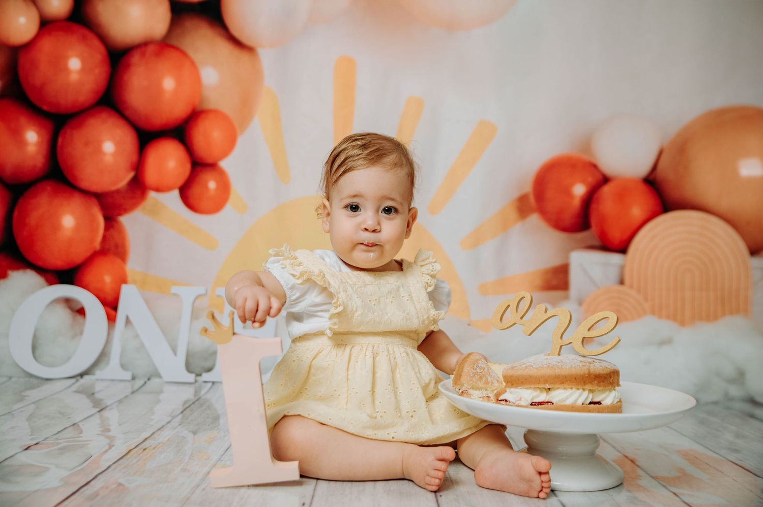 First birthday photo featuring baby in yellow dress reaching for cake with orange balloon decor.
