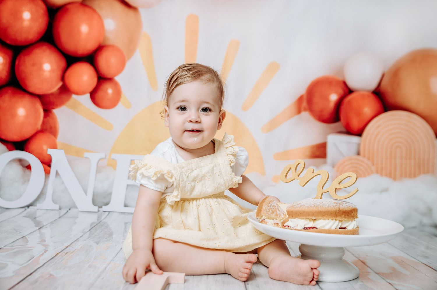 Smiling baby in cream dress poses for first birthday photos with cake and orange balloon backdrop.