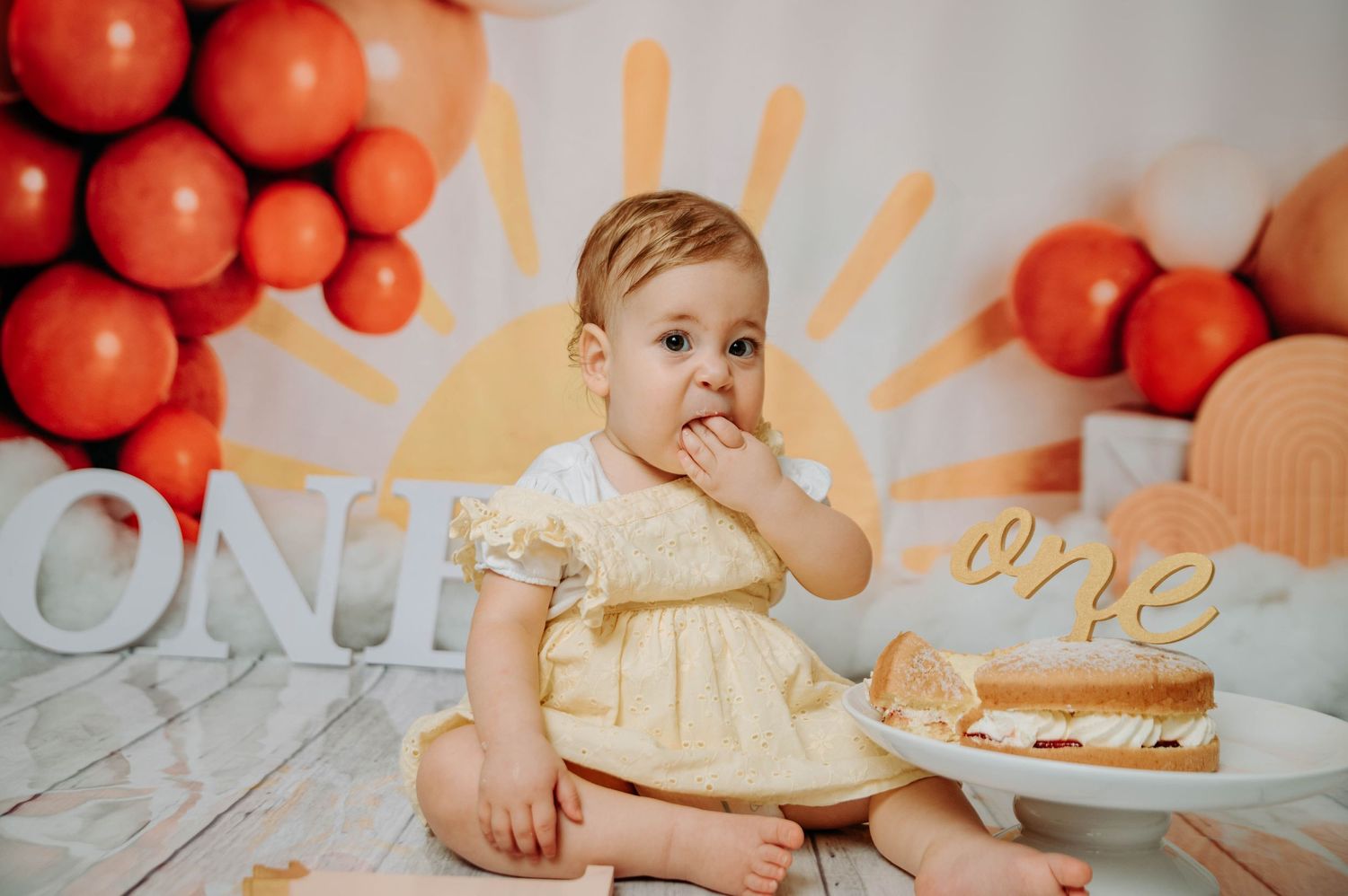 Baby in yellow dress touches face during first birthday photoshoot with cake and orange balloons.