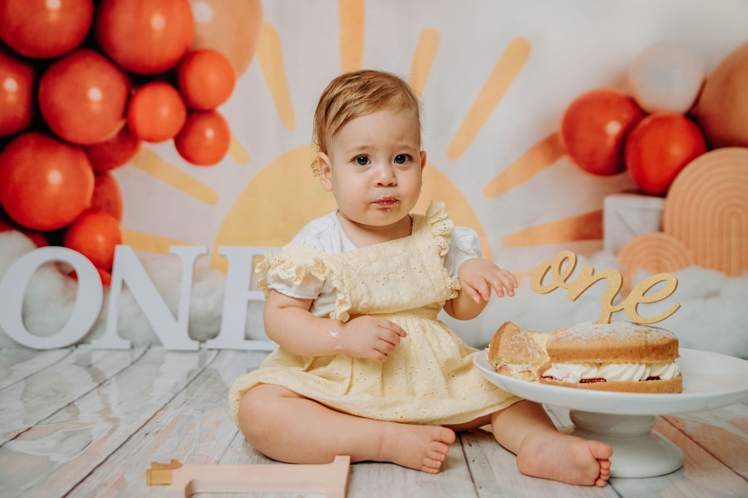 First birthday setup with baby in cream dress sitting near cake with orange balloon decorations.