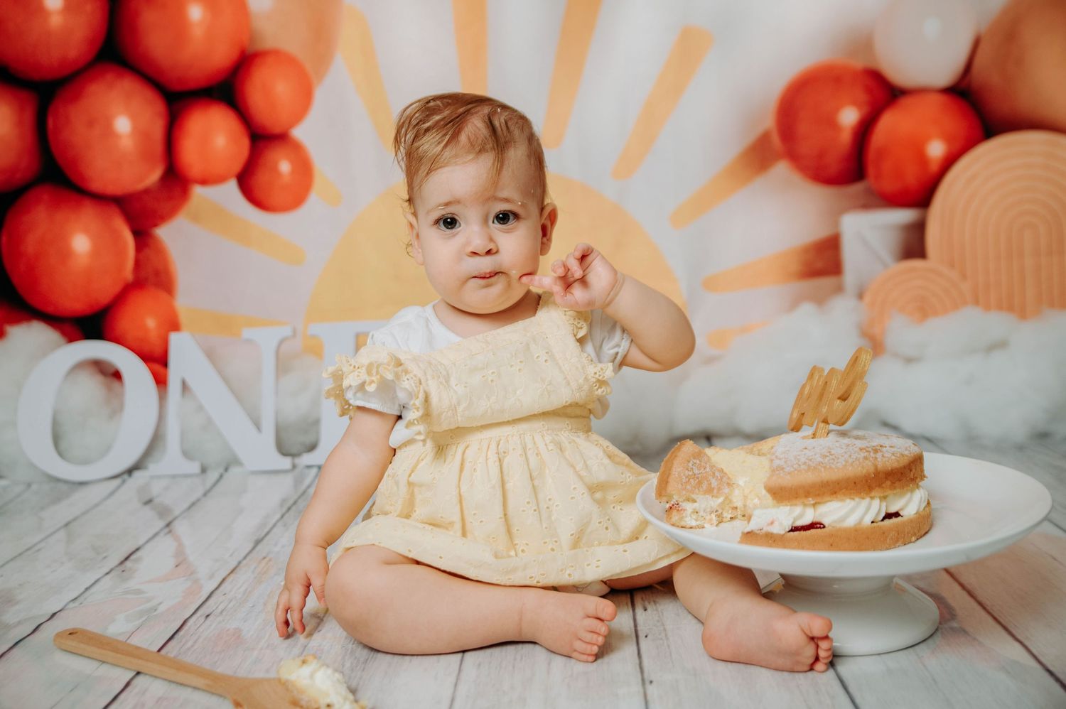 Baby in yellow dress reaches for birthday cake with orange balloon and sunburst background.