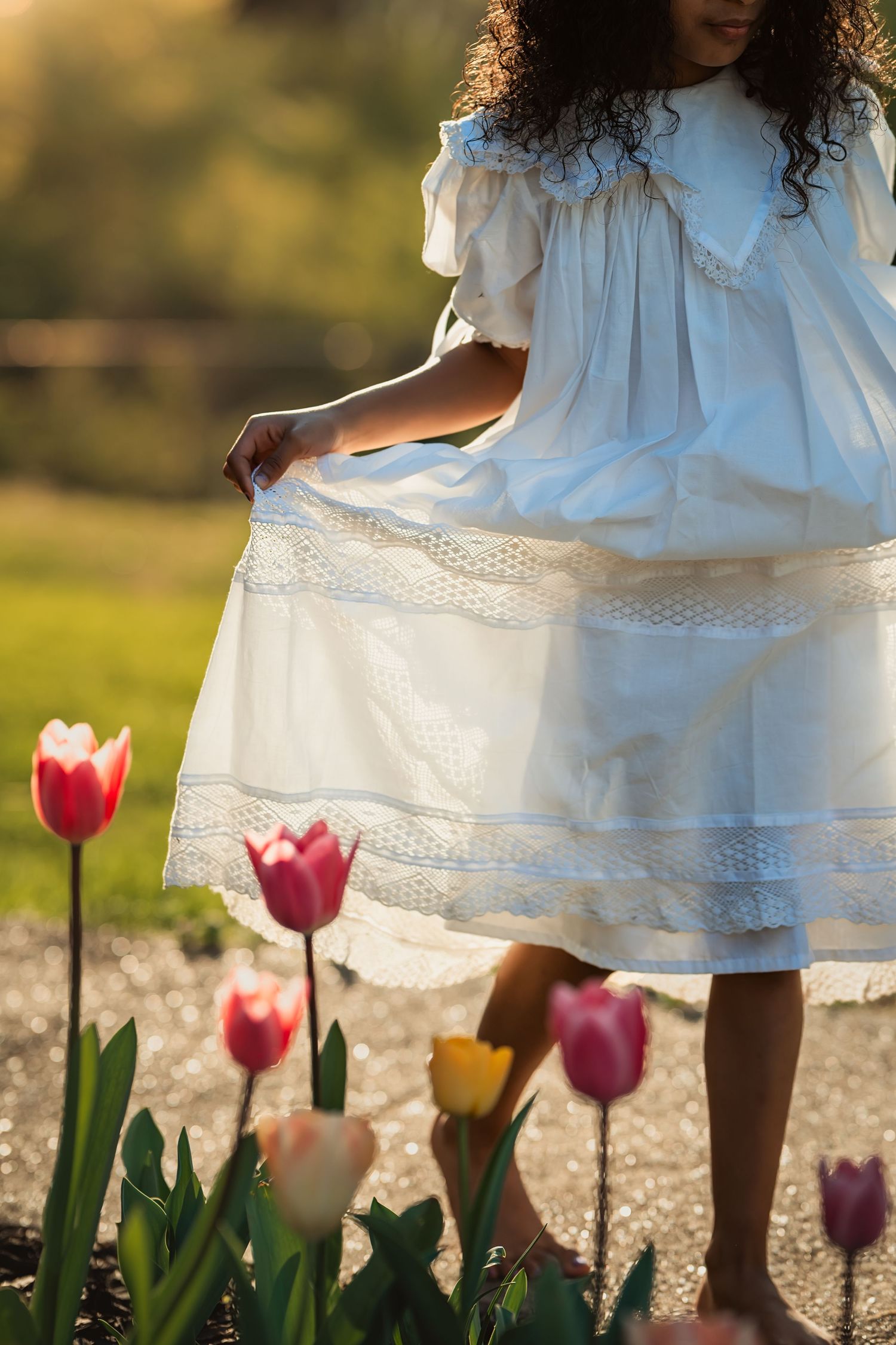 dark-skinned girl holds out white antique dress to catch the sun as she walks a path next to tulips at sunset