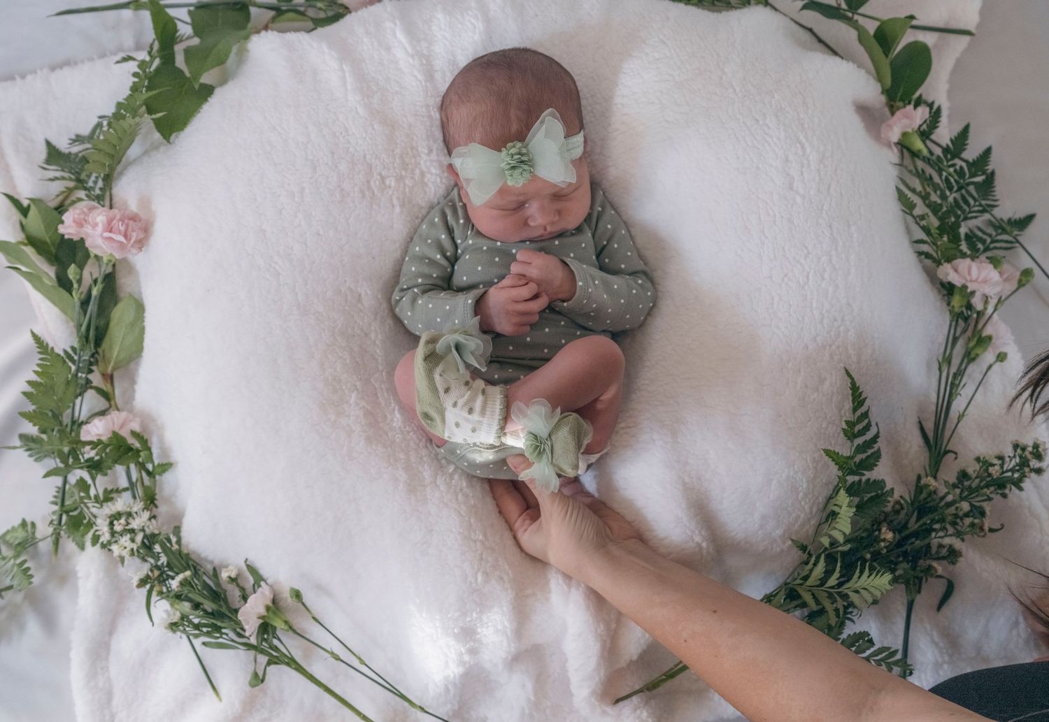 Newborn baby in green outfit with butterfly headband on white blanket with floral wreath and adult hand.