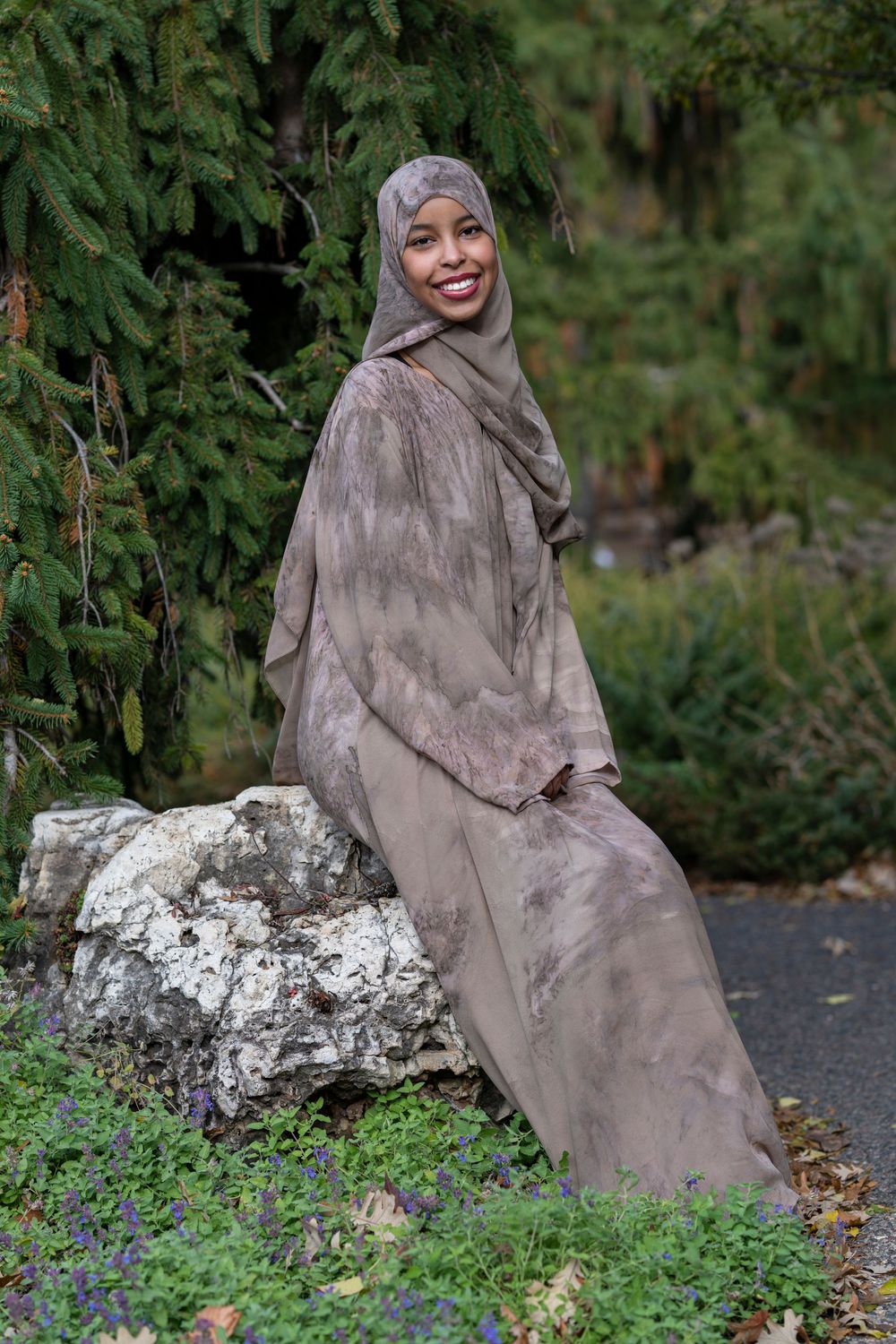 Smiling woman in a taupe hijab and abaya sitting on a rock in a lush green garden.