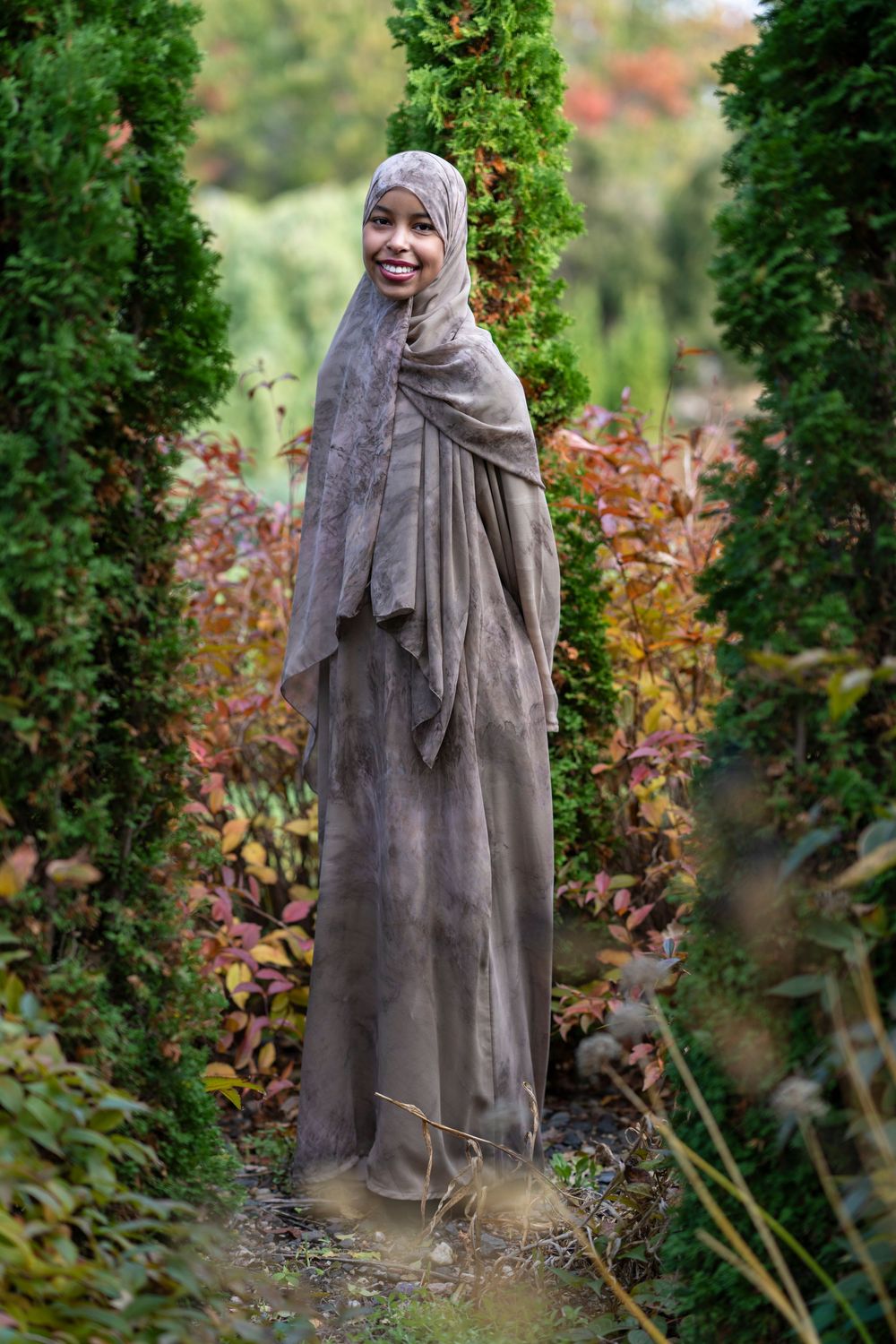 Smiling woman in a taupe hijab and abaya standing between tall green hedges in a garden.