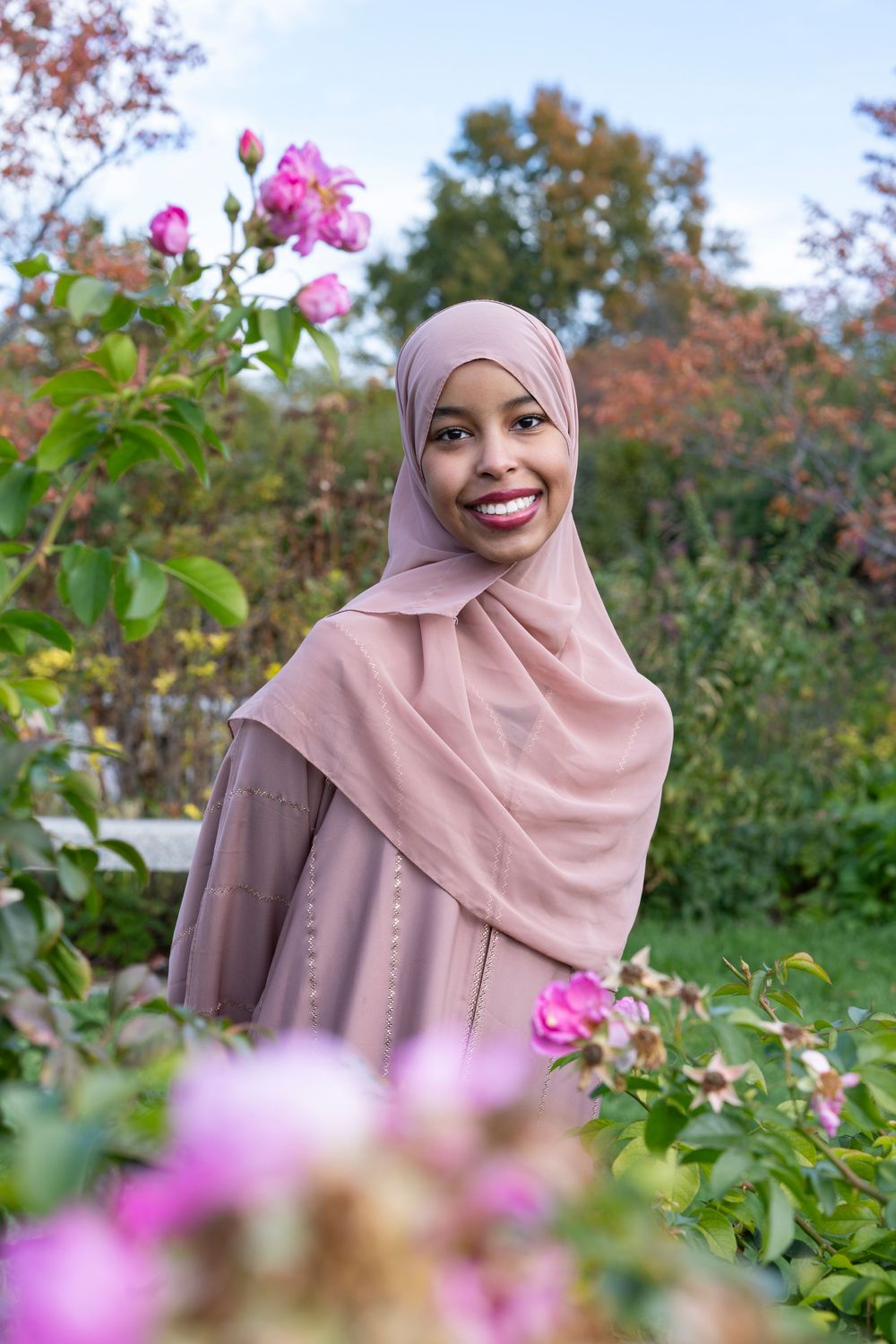 Smiling woman in a pink hijab and dress standing among blooming pink roses in a garden.