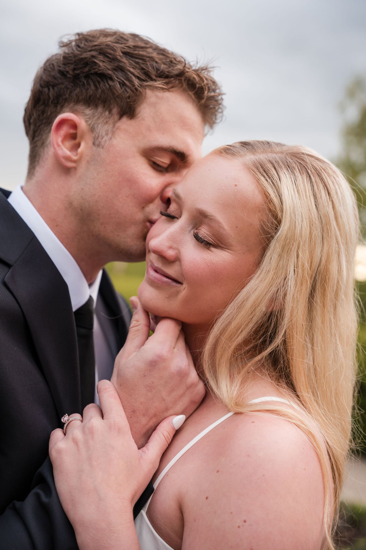A tender romantic moment between a couple on their wedding day, sharing a gentle kiss and embrace outdoors.