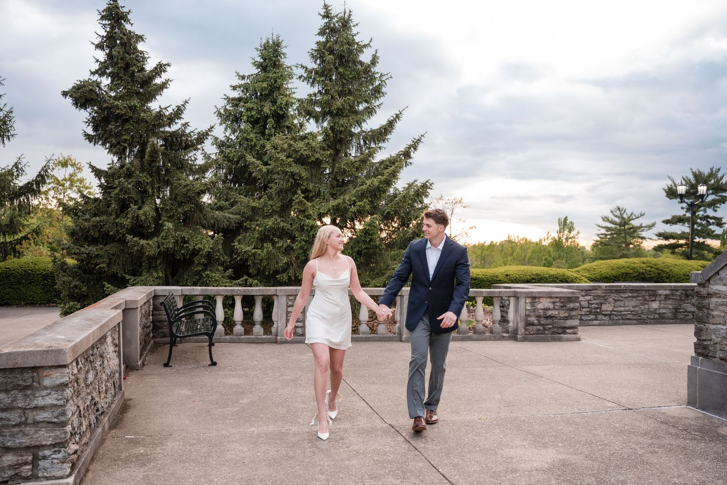 A couple walks hand in hand on a stone terrace with evergreen trees in the background on a cloudy day.