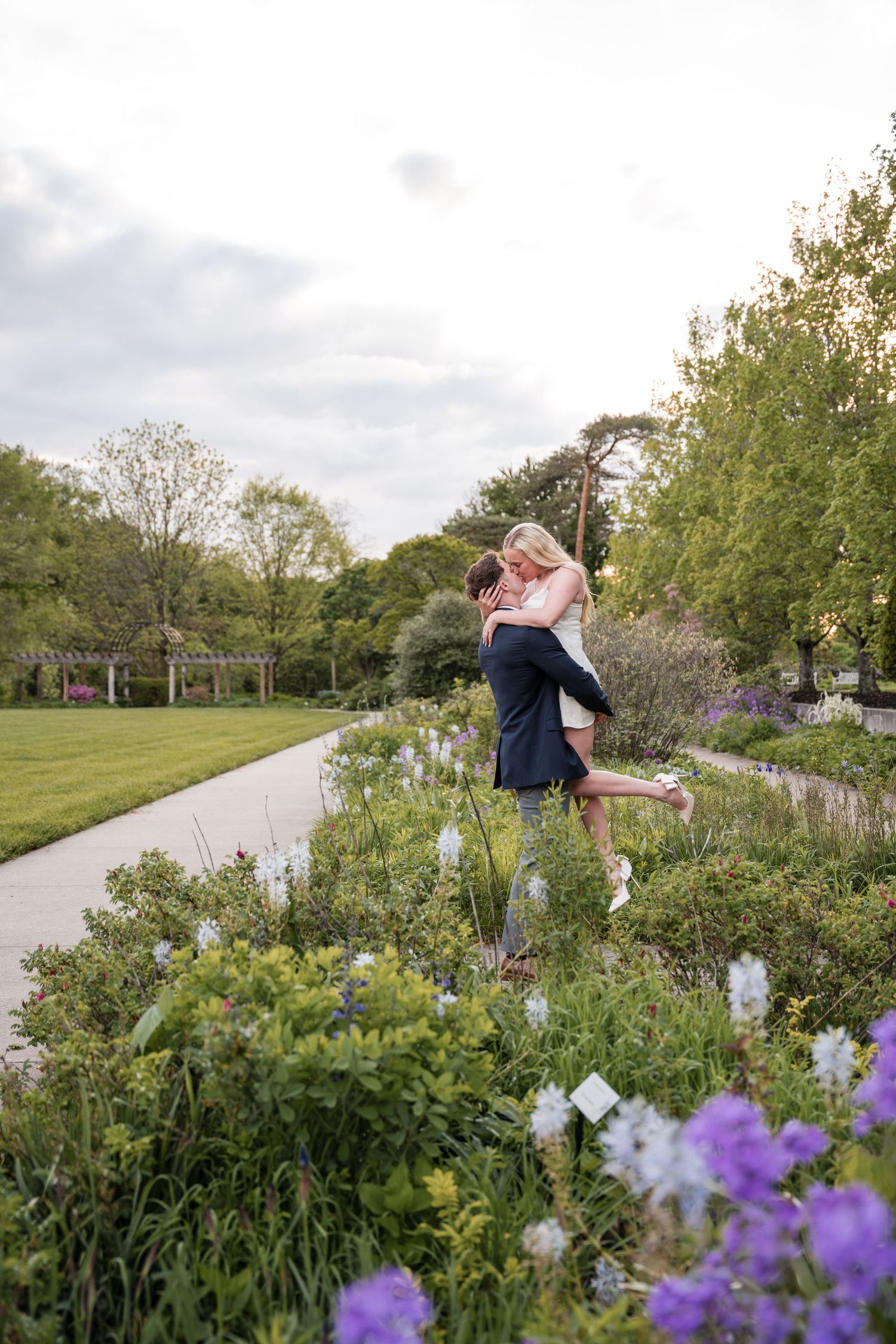 Two people sharing a romantic embrace along a garden path surrounded by blooming purple flowers and greenery.
