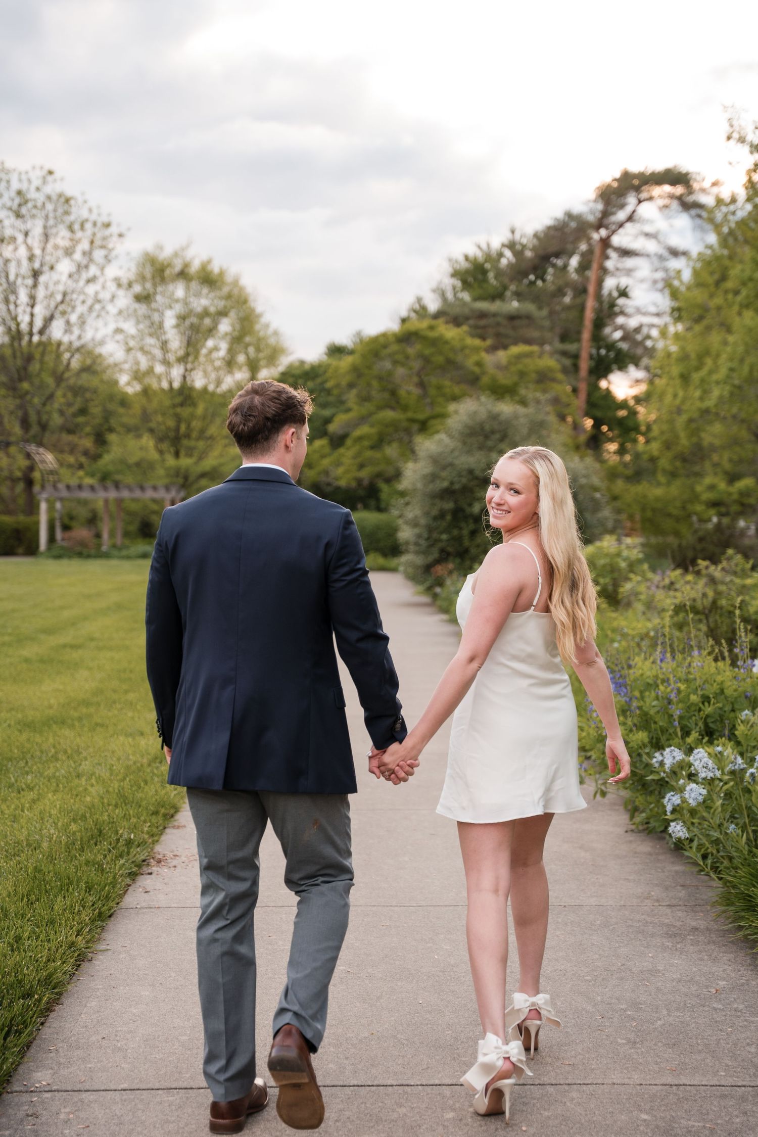 A couple walks hand in hand on a garden path with lush green landscaping and blooming flowers on a spring day.