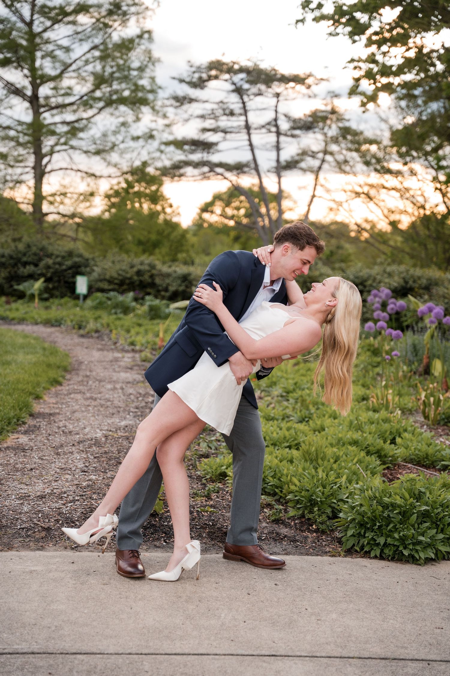 Romantic couple embrace and dip while dancing on garden path at sunset with blooming flowers in background.
