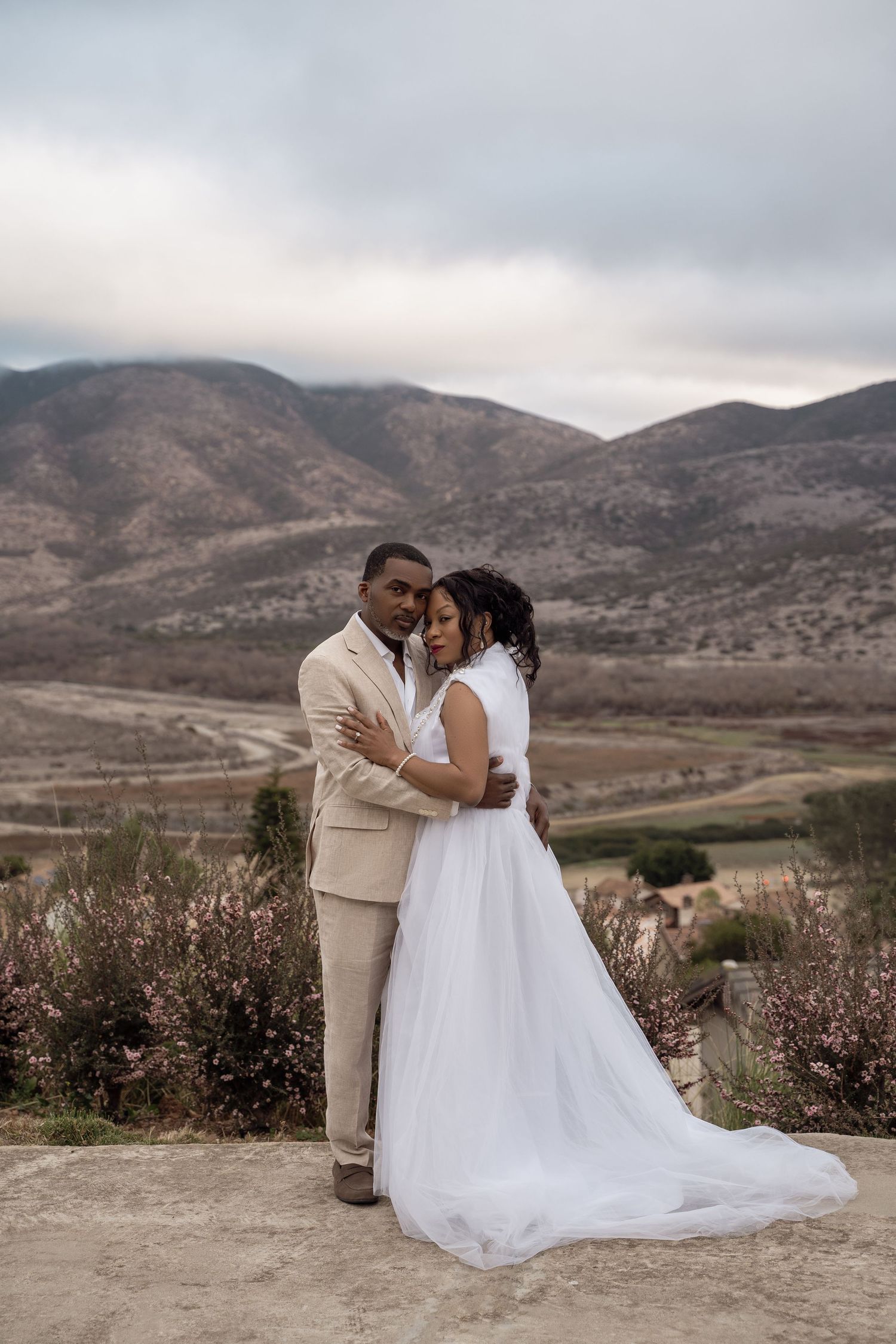 A couple shares an intimate moment in their wedding attire against a dramatic mountain landscape with cloudy skies.