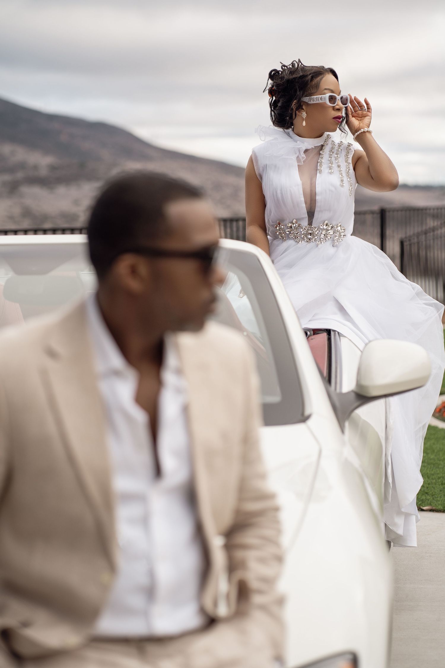 Stylish couple in elegant attire pose with a luxury white car against a mountainous backdrop on an overcast day.
