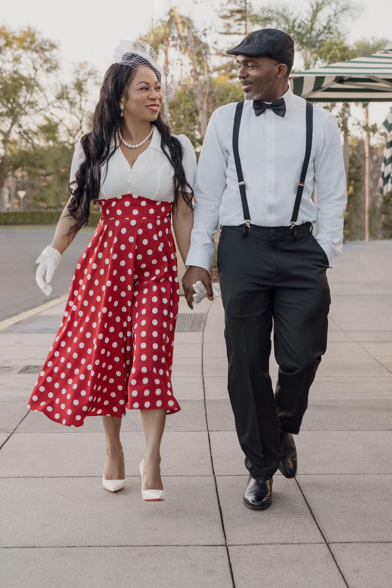 A couple in vintage-inspired outfits with polka dot dress and suspenders walk together on a paved outdoor path.