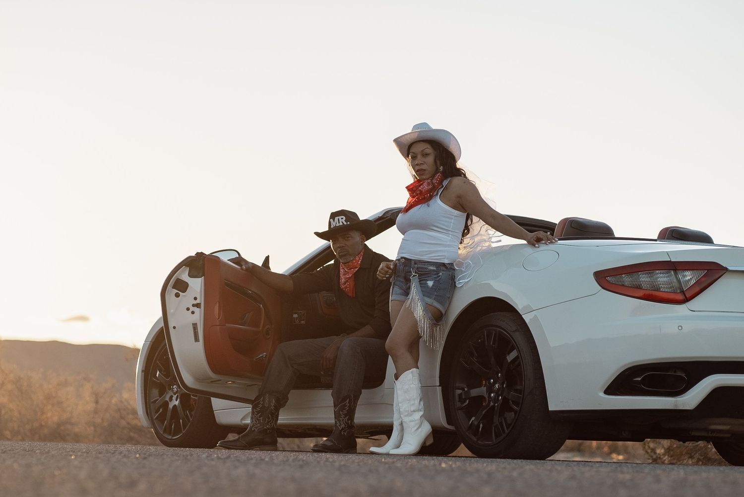 Two people in western attire lean against luxury sports cars on a desert road at sunset.