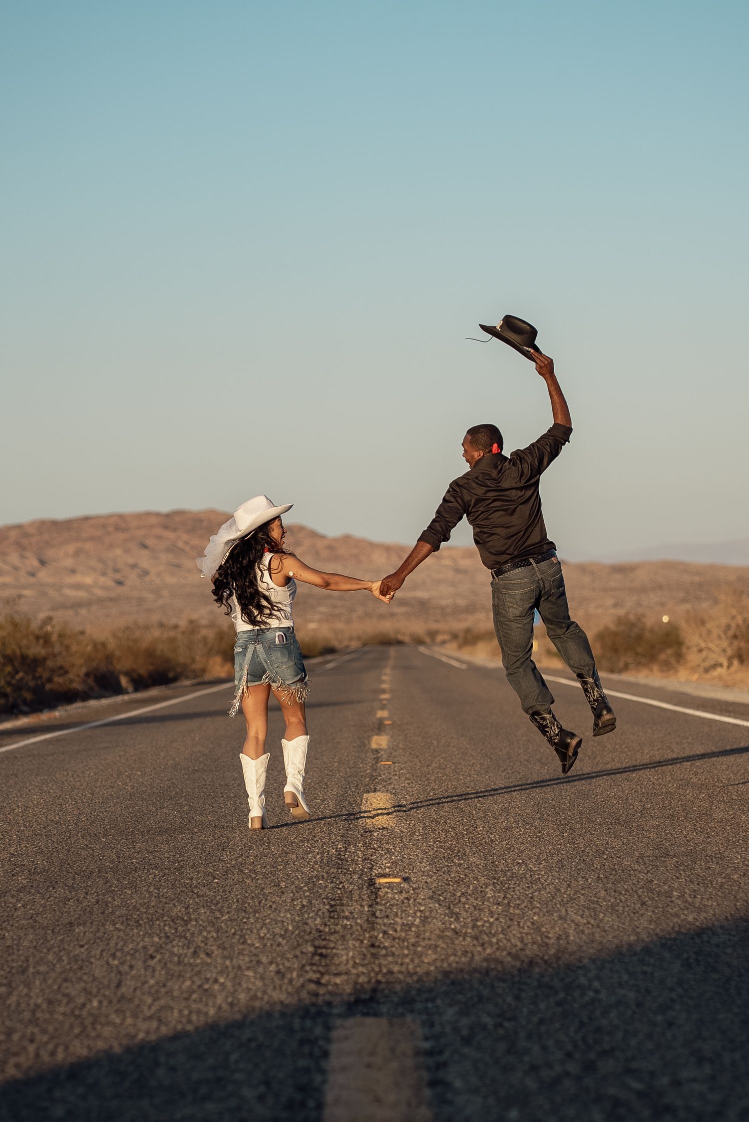 A couple in western attire dance and jump playfully on an empty desert highway at sunset.