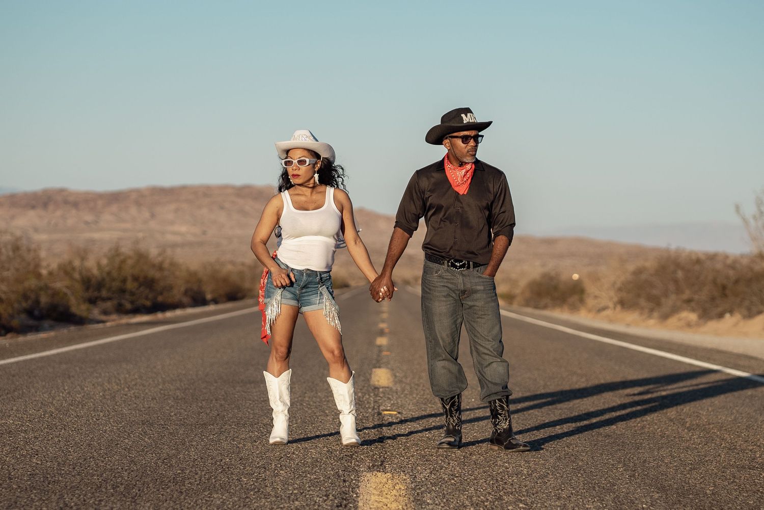 Two people in western style outfits holding hands while roller skating and walking on an empty desert highway.