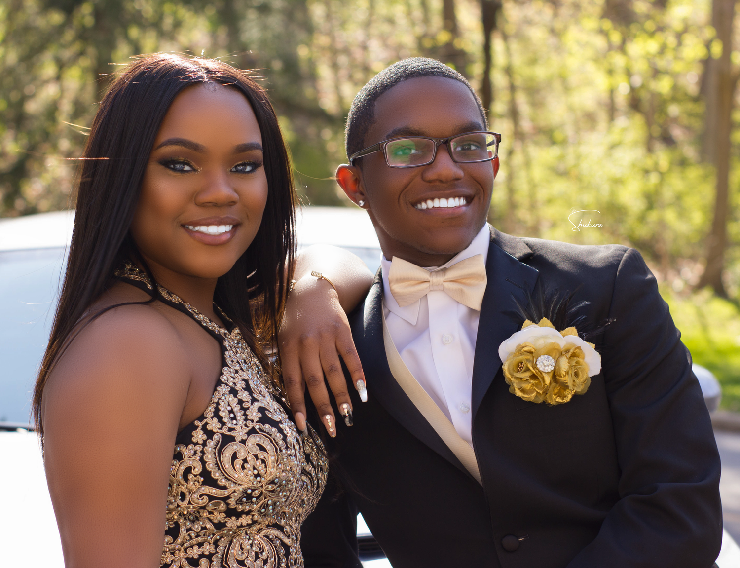 Formal couple poses outdoors wearing prom attire with gold accents.