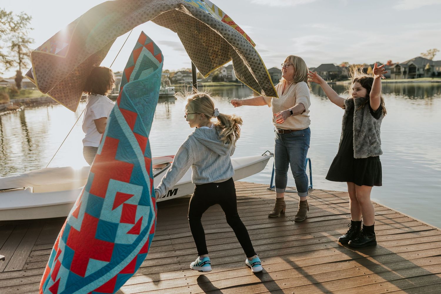 A woman laughs with her granddaughters as they throw handmade quilts in the air on a boat dock at golden hour.