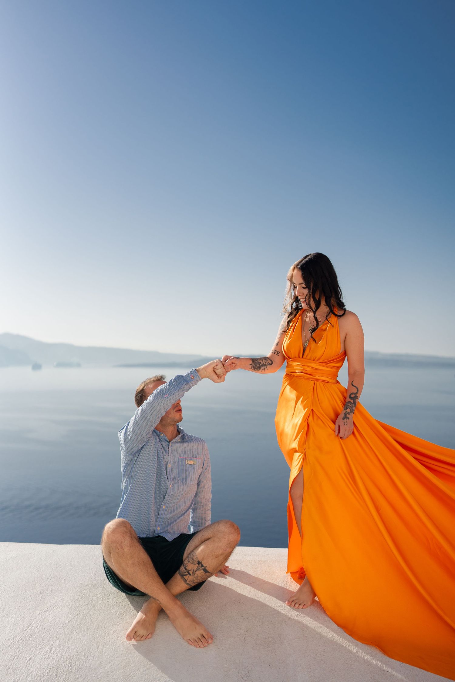 Couple in orange dress and casual wear pose on white rooftop overlooking Mediterranean Sea.