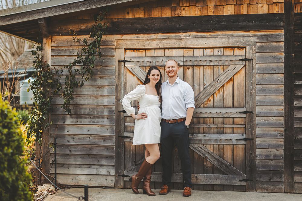 Couple in light colored outfits posing in front of rustic wooden barn door.