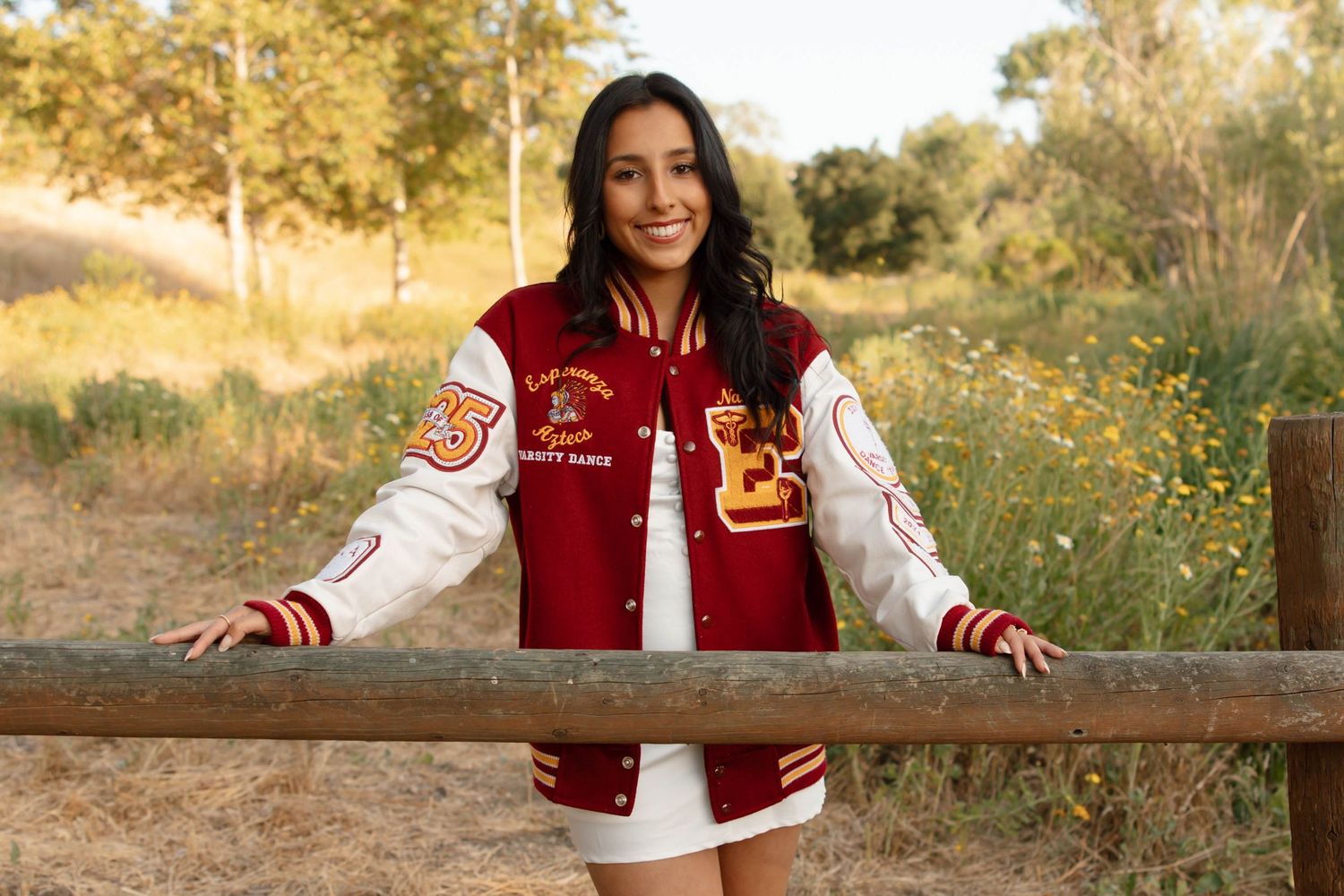 A person in a red and white varsity jacket poses by a rustic fence in a field for graduation and senior photos in Orange County