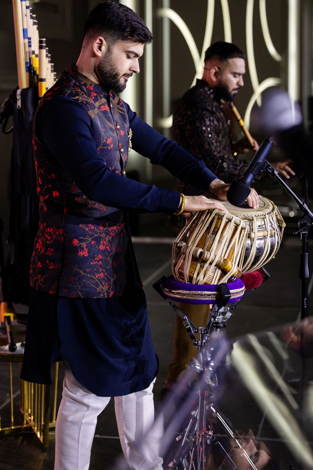Musician in navy and red floral jacket performs on traditional tabla drums during cultural performance.