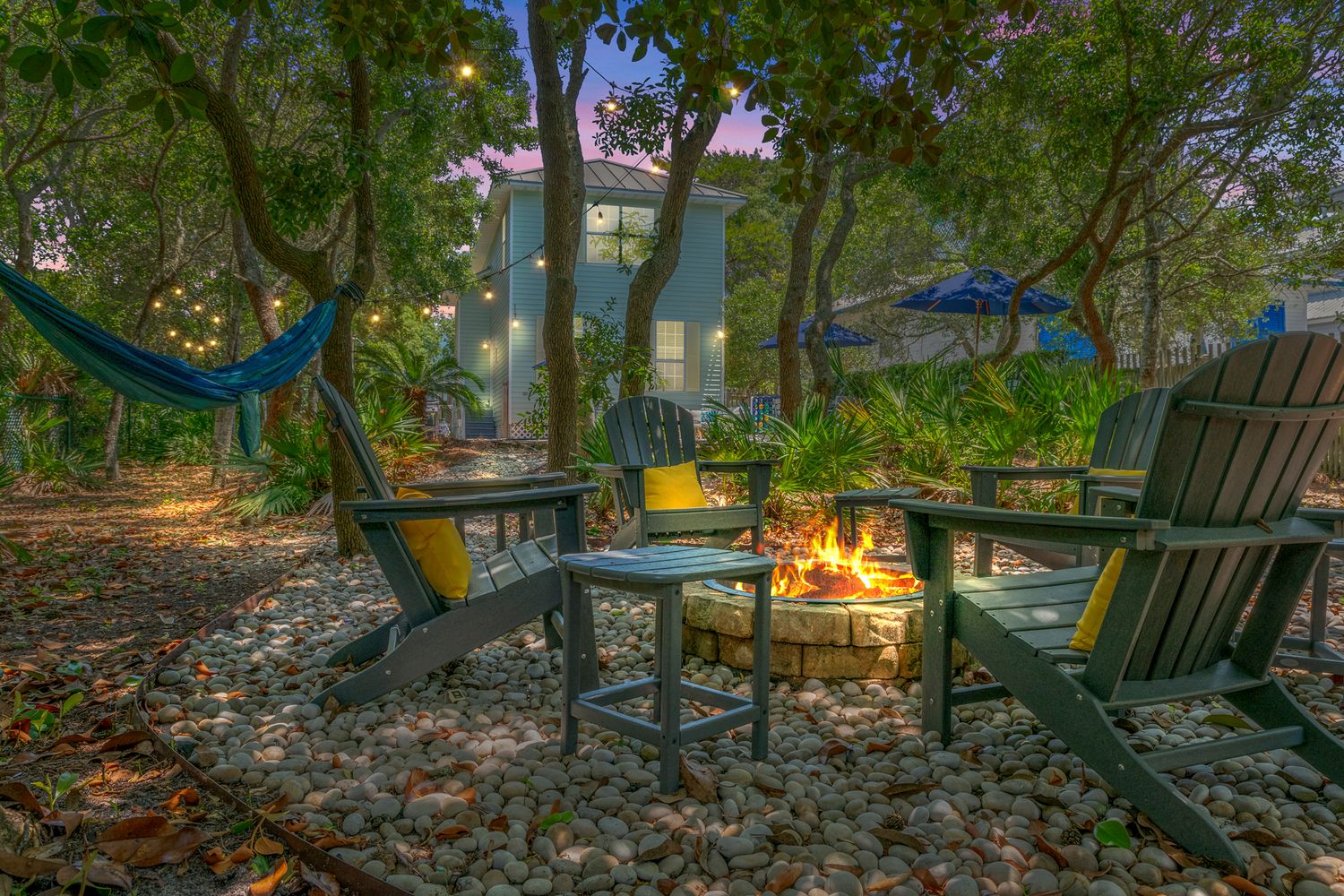 Cozy backyard fire pit area with Adirondack chairs and hanging hammocks among trees.