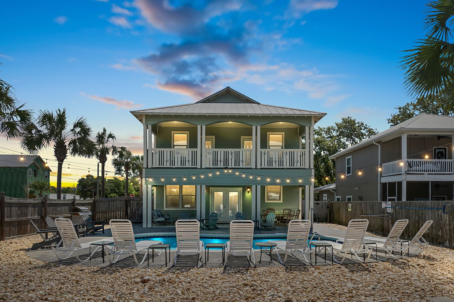 Two-story beach house with wraparound porch and pool illuminated at dusk.