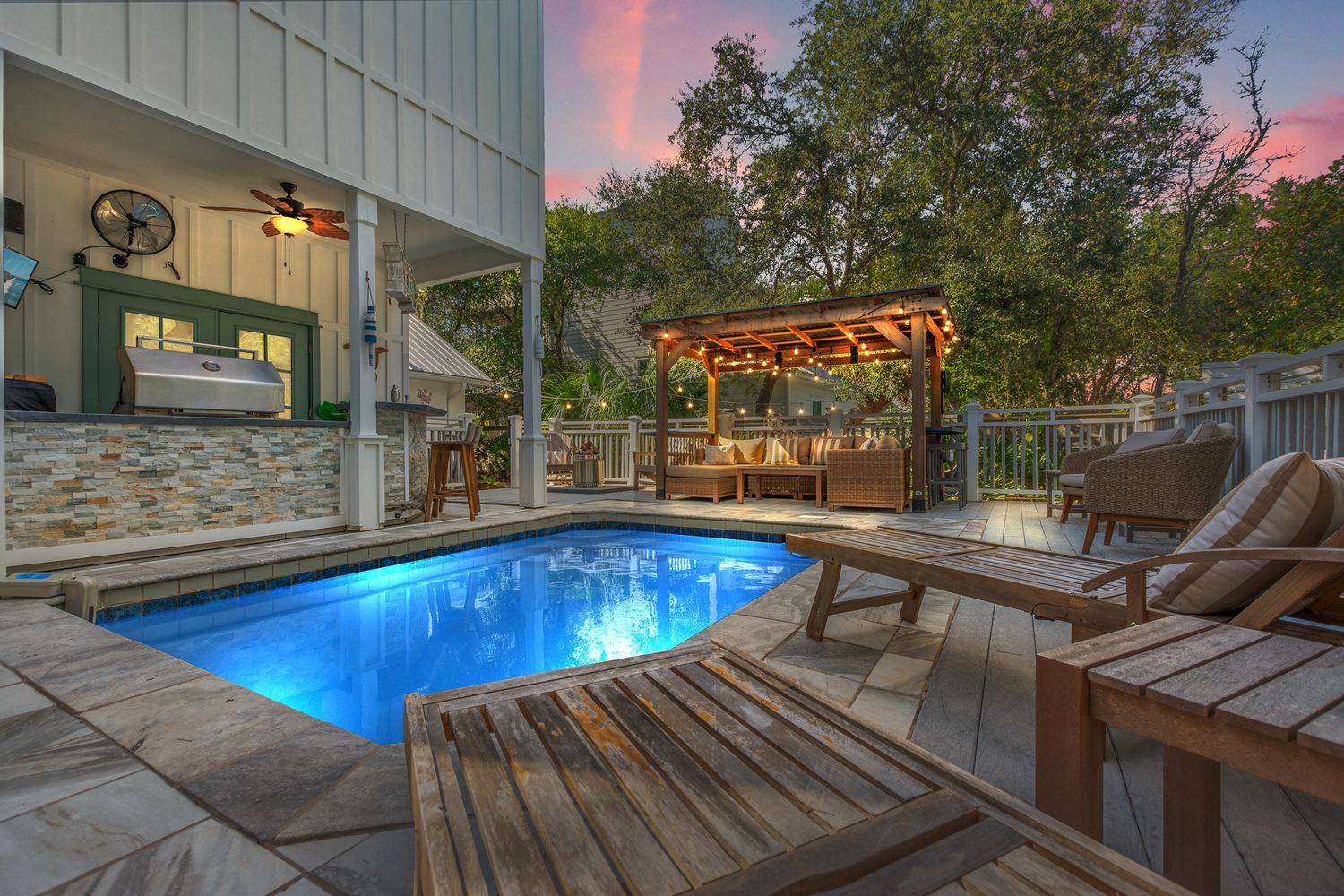 Illuminated backyard pool deck with wooden lounge chairs and covered pergola at twilight.