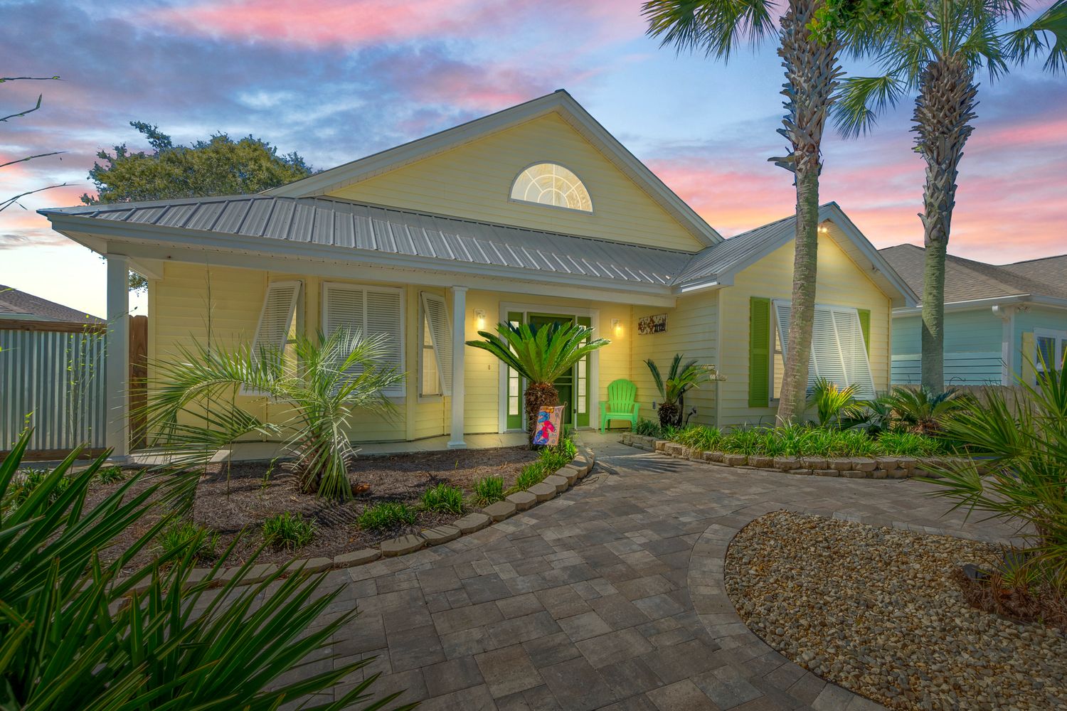 Coastal home with green metal roof and tropical landscaping at sunset with pink and purple sky.
