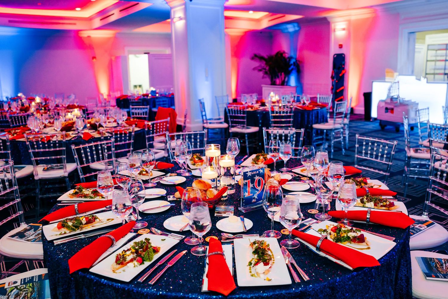 Elegant banquet hall with navy blue tablecloths, red napkins, crystal chairs, and colorful uplighting for a formal event.