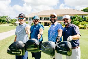 Four golfers smiling and extending their golf clubs toward the camera on a sunny tropical golf course.