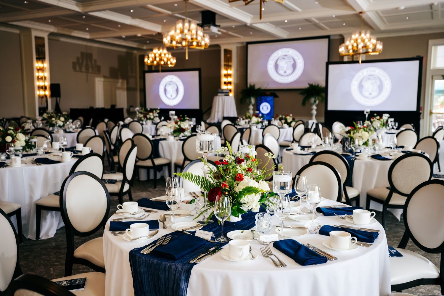 Elegant banquet hall with round tables, navy blue napkins, floral centerpieces, and presidential seal screens.
