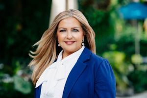 Professional wearing navy blazer and white blouse poses against blurred green background.