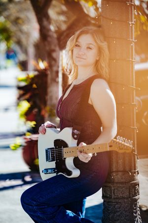 Musician poses with white electric guitar in outdoor sunlit setting with string lights.