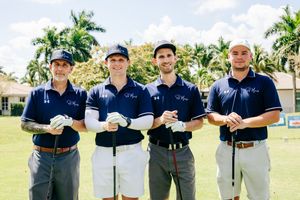 Four men in matching navy blue polo shirts pose with golf clubs on a lush tropical golf course.